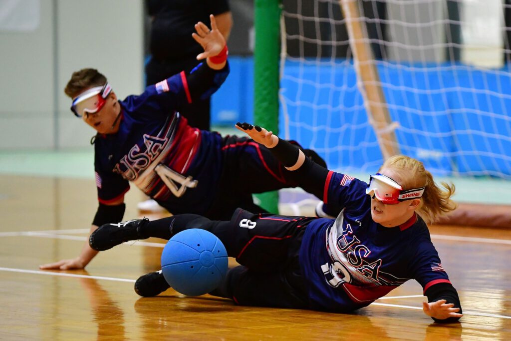 two people with white glasses laying on gym floor sprawled out in front of heavy blue wall blocking from goal 
